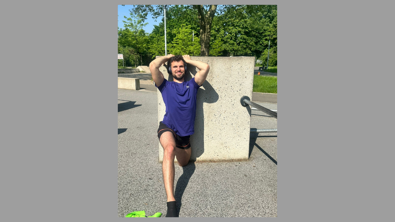 Man in a blue shirt and black shorts stretching outdoors during a morning workout. He is leaning against a concrete structure, enjoying the sunny weather in a park-like setting.