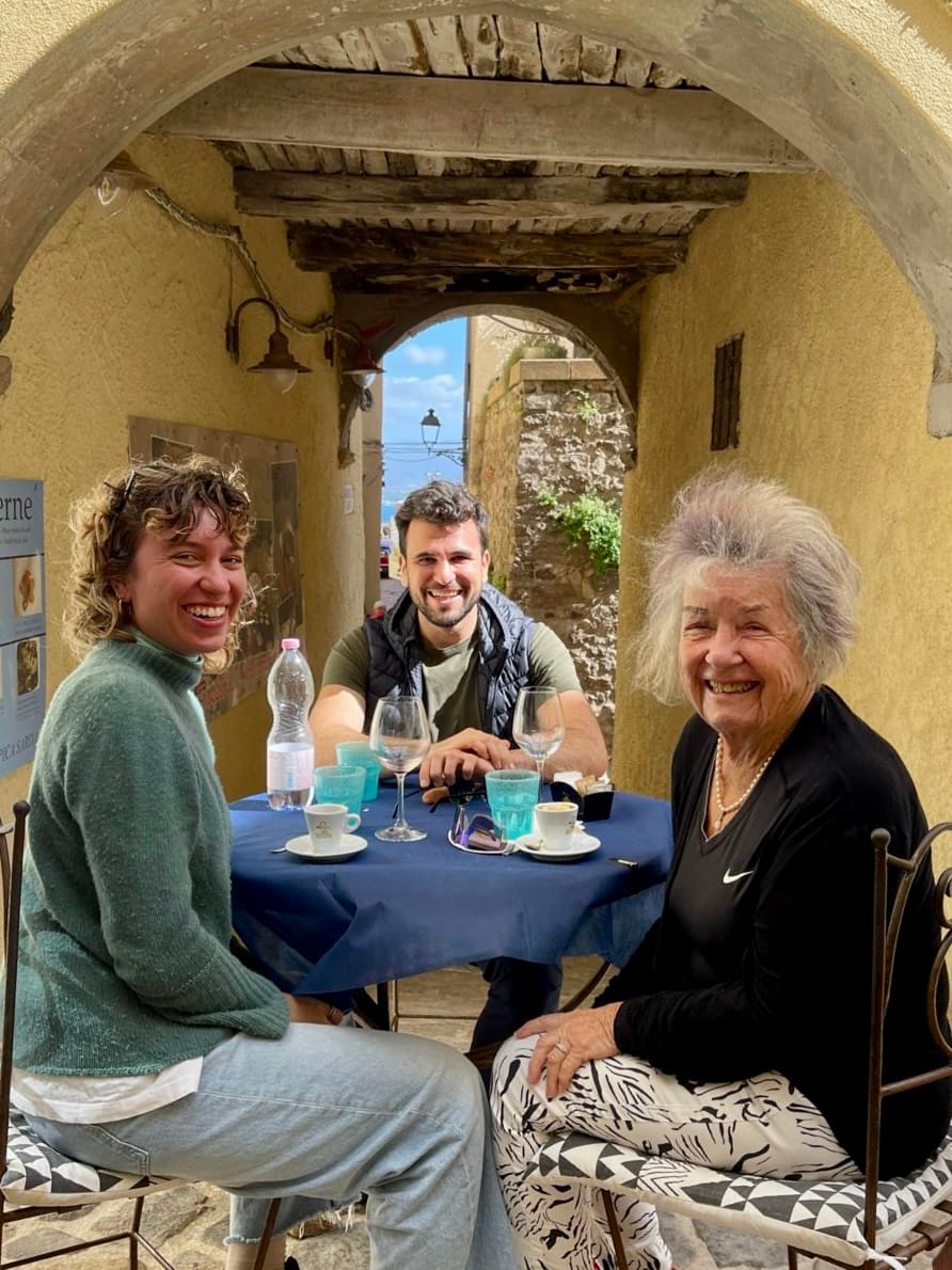 Three people are sitting at a table in a cozy alleyway under an old, rustic archway. On the left is a woman with curly hair wearing a green sweater, in the middle is a man with a beard and a vest, and on the right is an older woman with gray hair, smiling at the camera. The table has empty wine glasses, espresso cups, and a water bottle on it. The atmosphere is relaxed and friendly, with a Mediterranean vibe in the background.