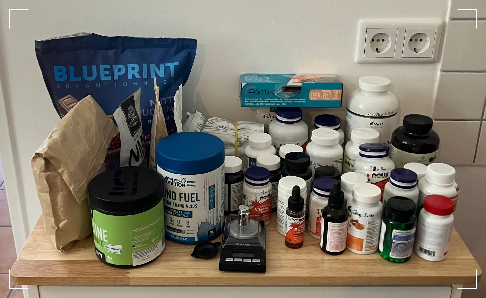 In the image, there is a wooden surface with a variety of supplements and health-related products neatly arranged. On the left, there’s a large blue bag labeled "Blueprint Bryan Johnson" and some other smaller bags. In the middle, there are containers labeled "Amino Fuel" and other supplements like powdered products. On the right, there is a cluster of various supplement bottles, including several from brands like NOW and Nu U Nutrition. Some bottles have dropper lids, indicating they are liquid supplements or tinctures. Also, there’s a small black device in the middle, which appears to be a scale or measuring tool. The products seem to be part of a detailed supplement routine, possibly inspired by Bryan Johnson's Blueprint regimen.