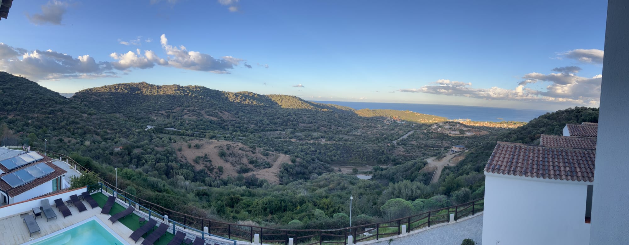 A panoramic view of a hilly landscape with lush greenery and scattered homes, stretching towards the horizon where the sea and sky meet. In the foreground, there is a pool area with lounge chairs, surrounded by a fence. The scene is bathed in soft light, with a few scattered clouds in the blue sky.