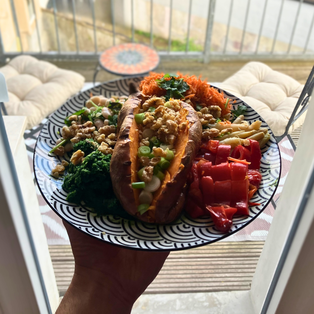 A balanced vegetarian meal served on a patterned black and white plate, held in a person's hand. The plate features a stuffed sweet potato topped with green onions and walnuts, alongside servings of shredded carrots, kale, chopped red peppers, and a small portion of asparagus. The background shows a balcony view with a table and a cushioned chair.