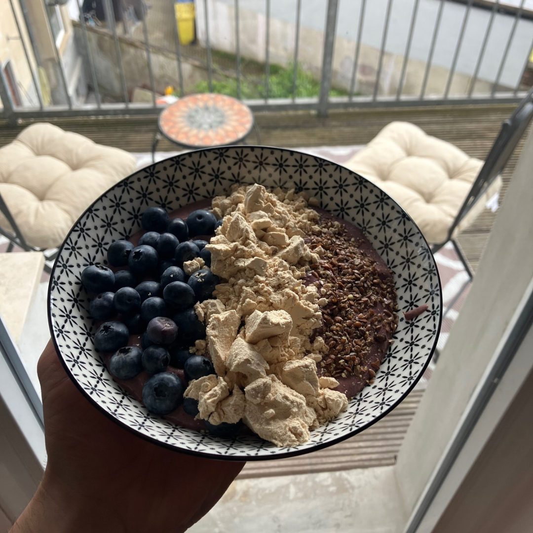 The Nutty Pudding with the topping: fresh blueberries, vanilla-flavored pea protein (30 grams), crushed linseed The bowl is patterned with black and white stars, adding a stylish touch. The setting includes a balcony view with a decorative table and a plush cushioned chair in the background.