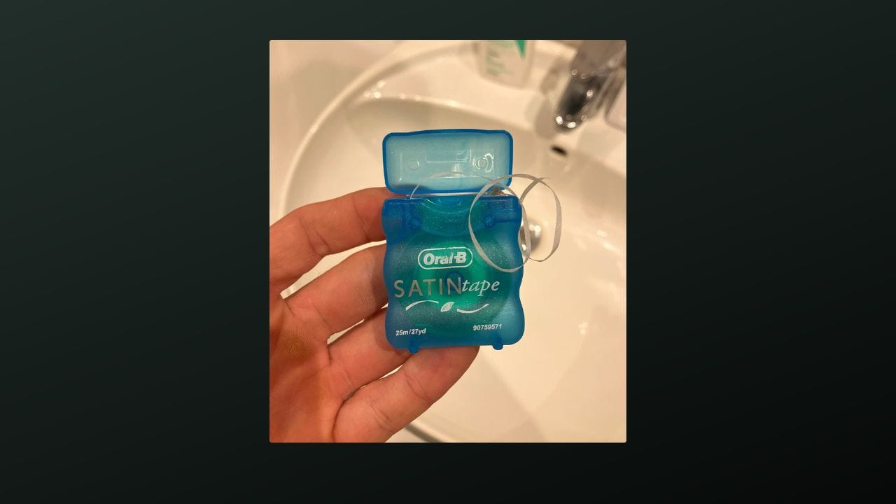 A hand holding a container of Oral-B SATINtape dental floss in a bathroom setting. The background shows a white sink and various toiletries, including a soap dispenser and a lotion bottle on the counter.