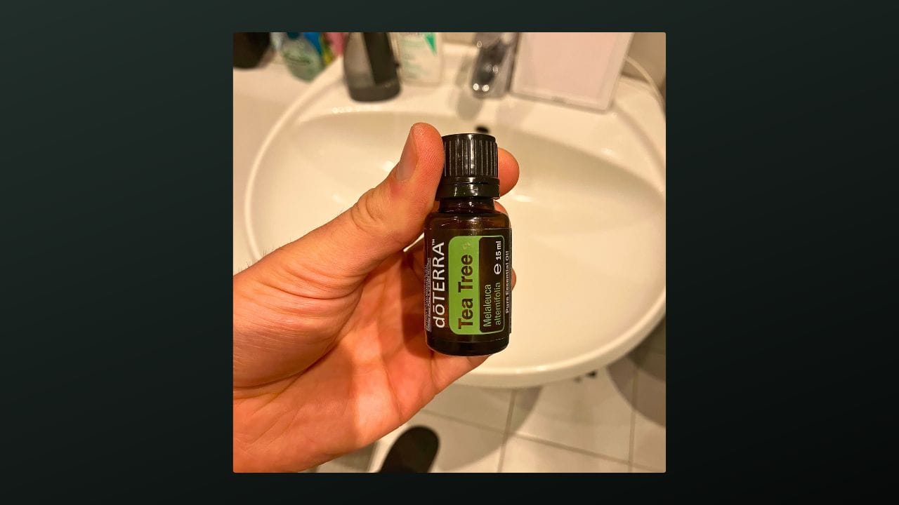 A hand holding a small brown bottle labeled "doTERRA Tea Tree Essential Oil" in a bathroom setting. The background shows a white sink and various toiletries on a counter.