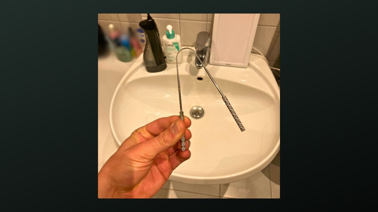 A hand holding a metal tongue scraper in a bathroom setting. The background shows a white sink and various toiletries, including a black soap dispenser, a white container, and other items on a counter.